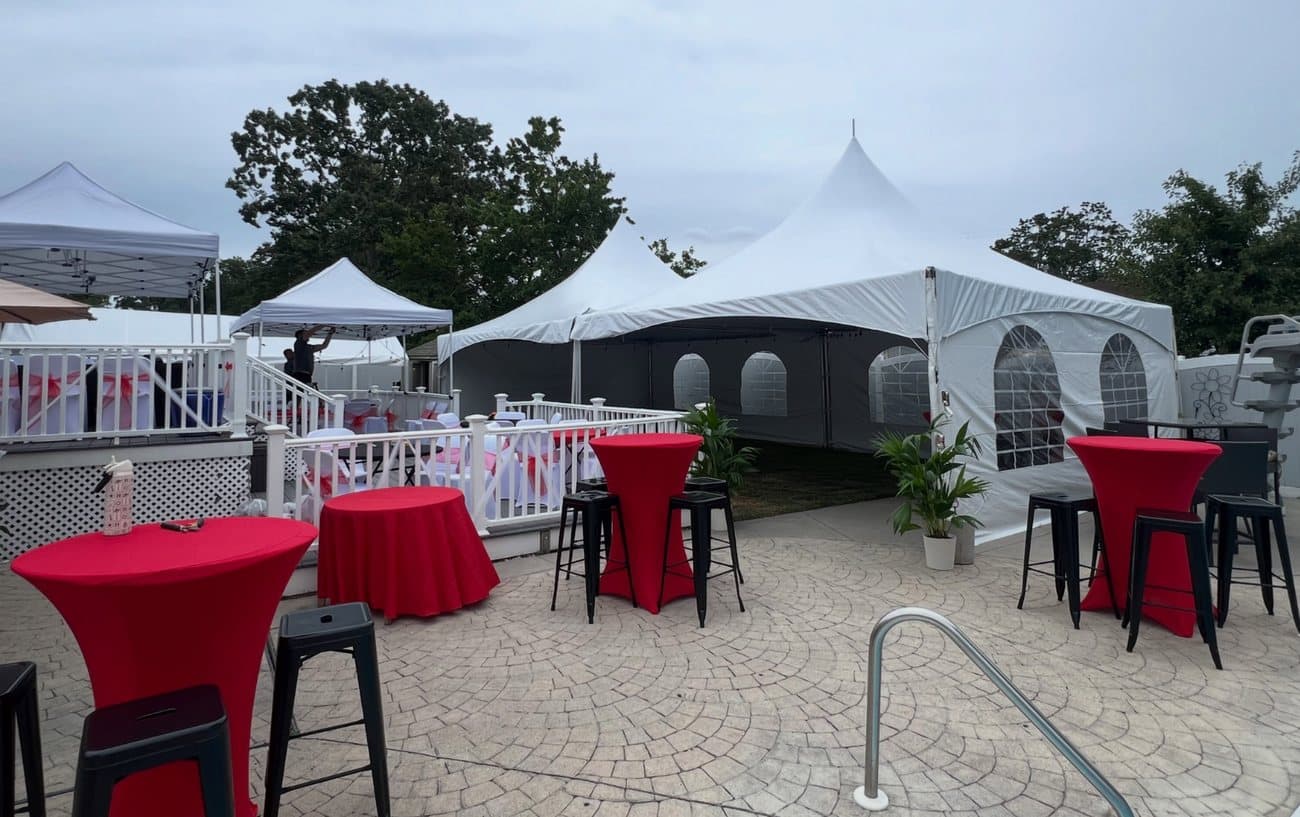 A high peak tent with black sidewalls and arched cathedral windows on a paver patio, set with red-linened cocktail tables and black stools.