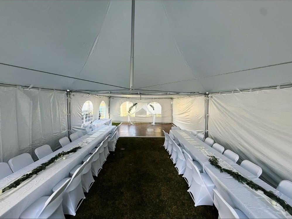 The inside of a tent with long banquet tables in white linens, white chair covers on every chair and greenery runners down the tables.