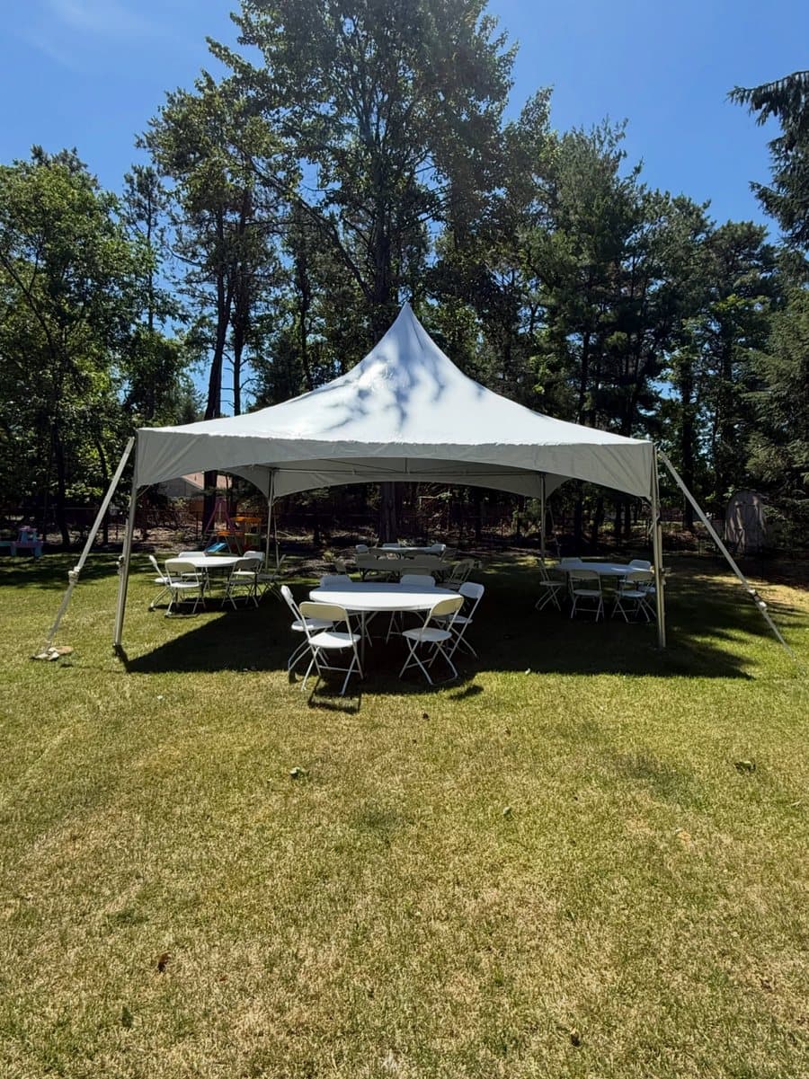 A 20 by 20 high peak tent in a pine-shaded backyard with a 60 inch round table and white folding chairs underneath.
