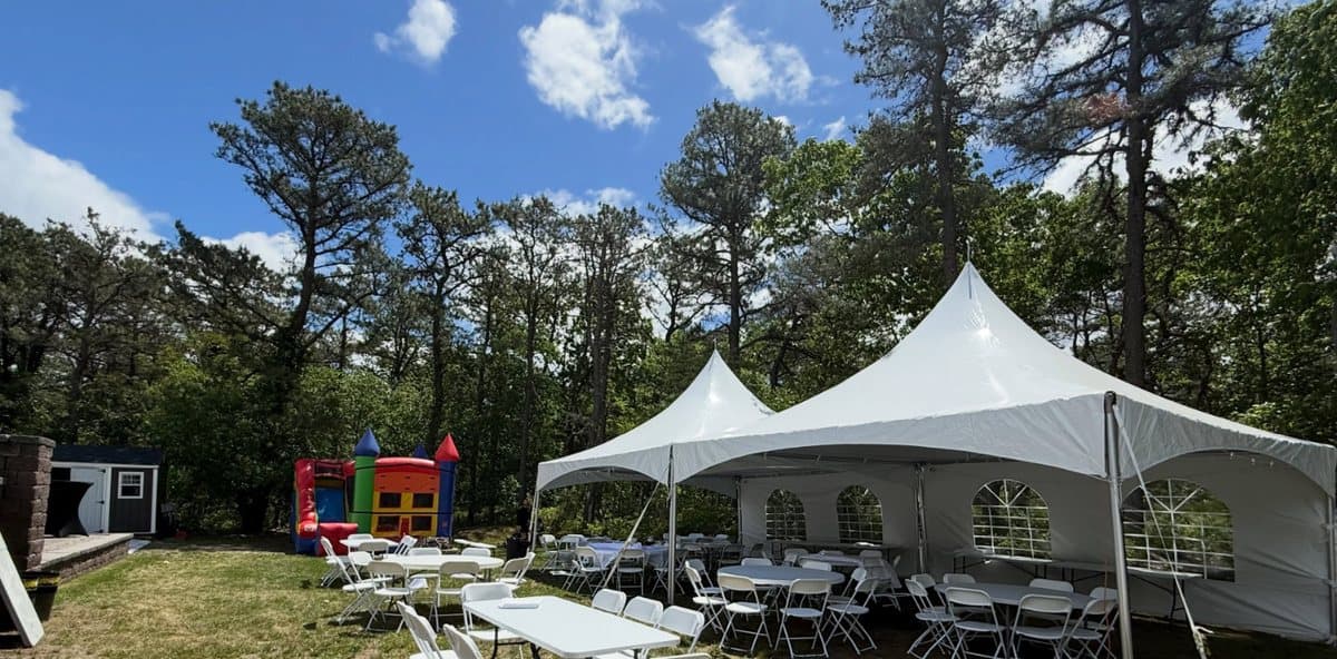 A 20 by 40 twin peak tent with white sidewalls and arched windows, tables and white folding chairs set up on a wooded backyard lawn.